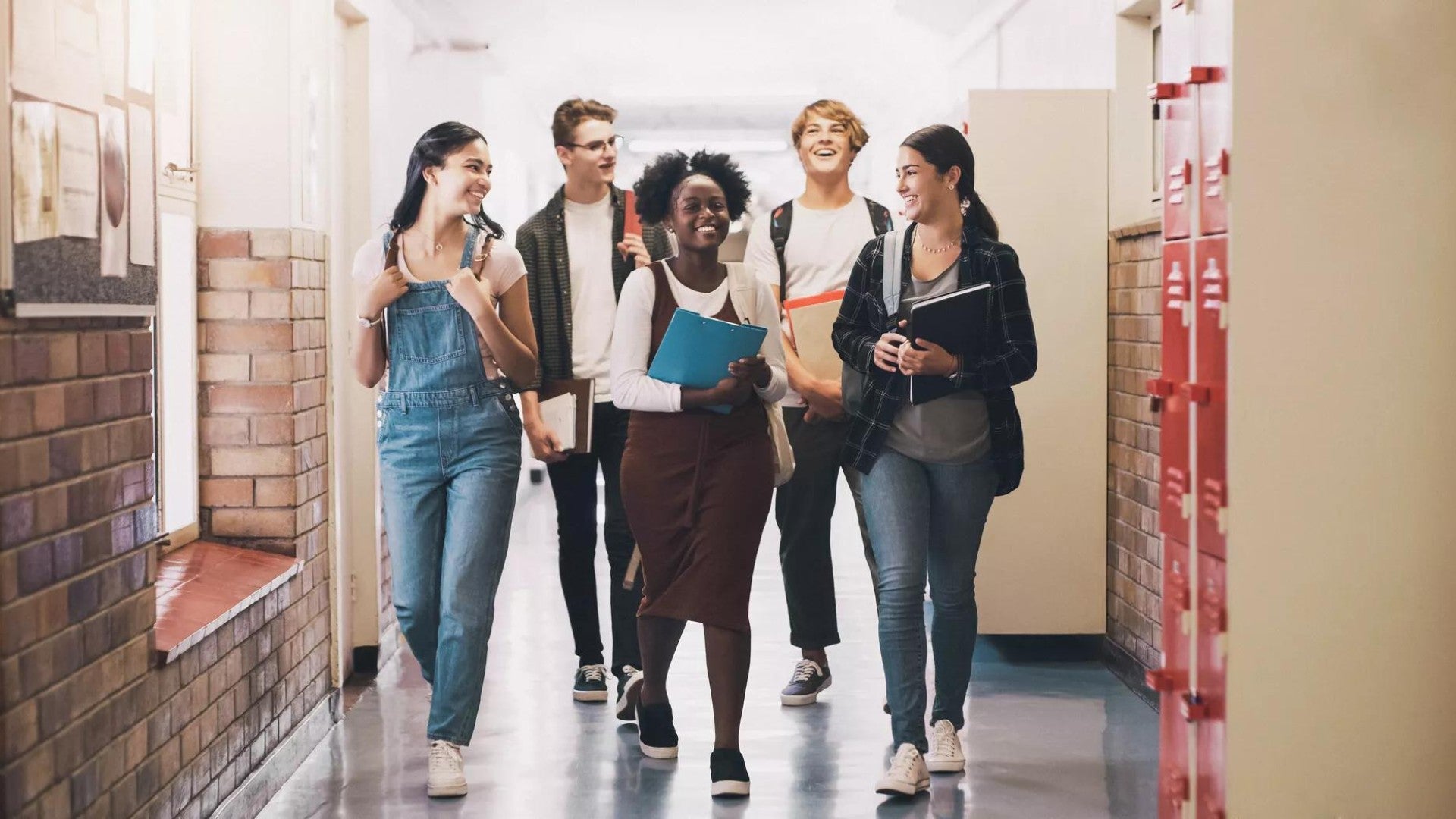 Five high school teenagers (two boys, three girls) carry books and backpacks as they walk down the hallway at school.