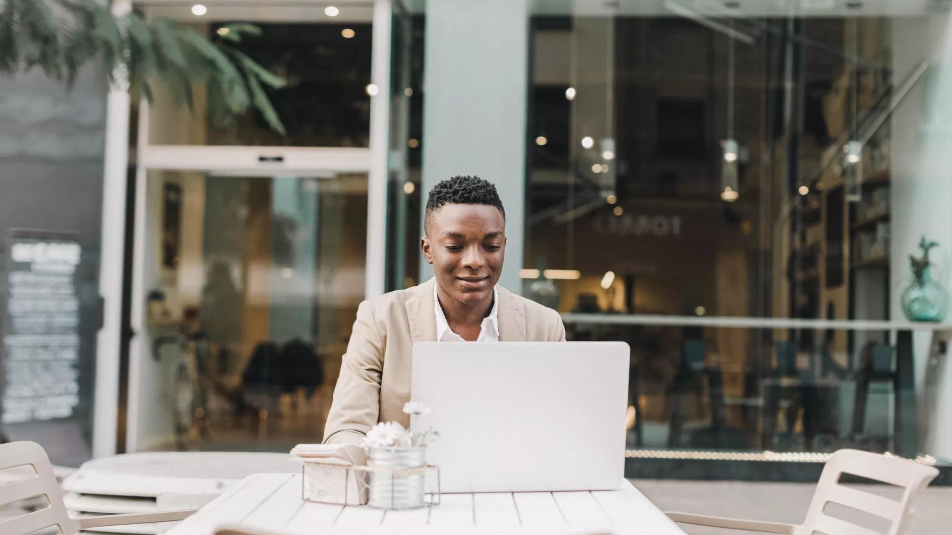 Young man working on his laptop at a patio table outside of a business.