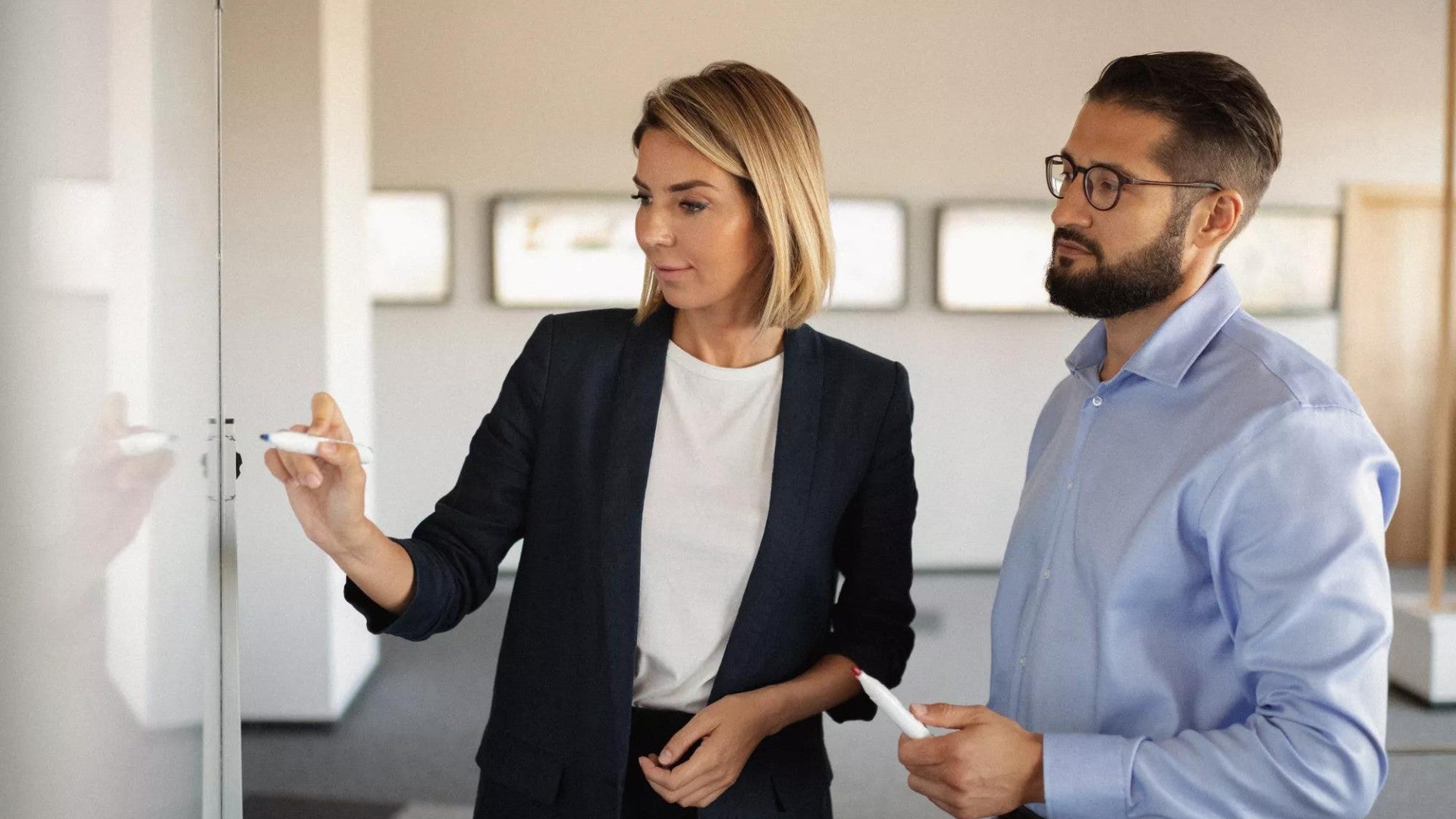 Two employees, a woman and a man, looking at and writing on a whiteboard in the office.