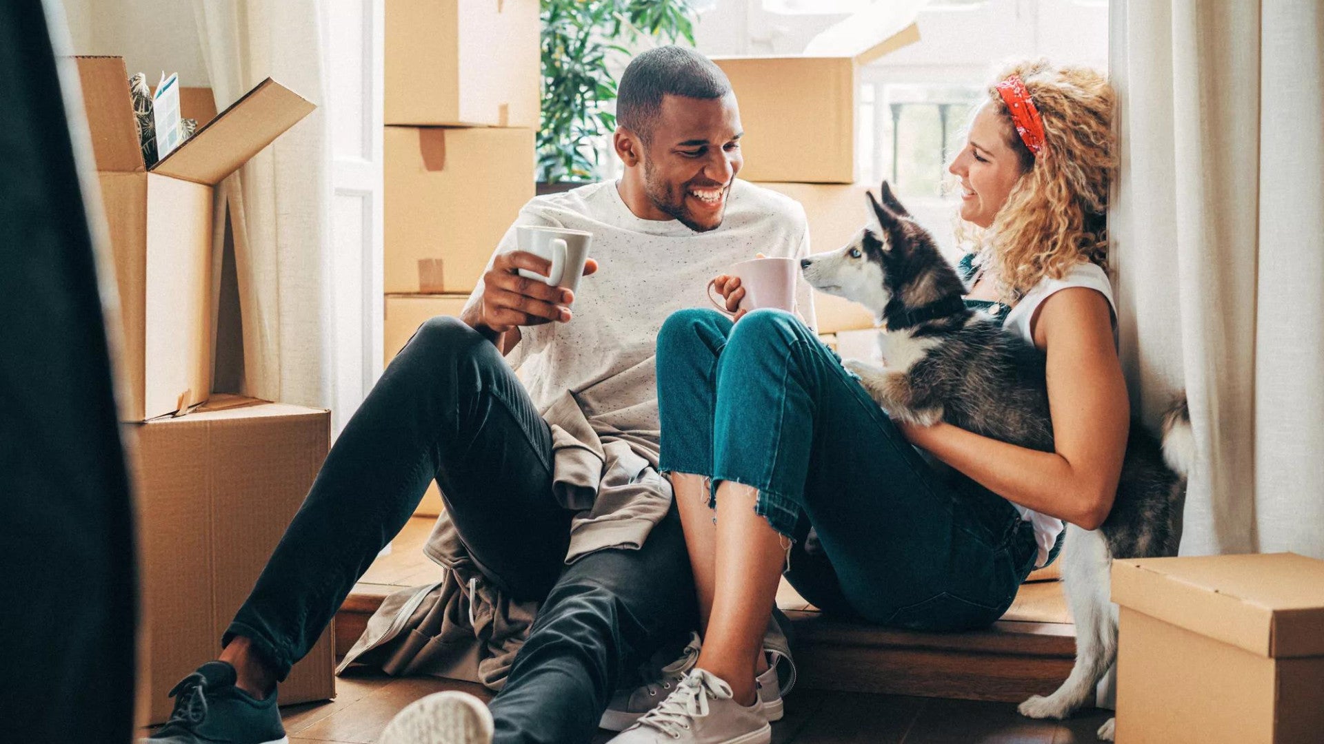 Man and woman sit on floor of home holding dog and surrounded by moving boxes.