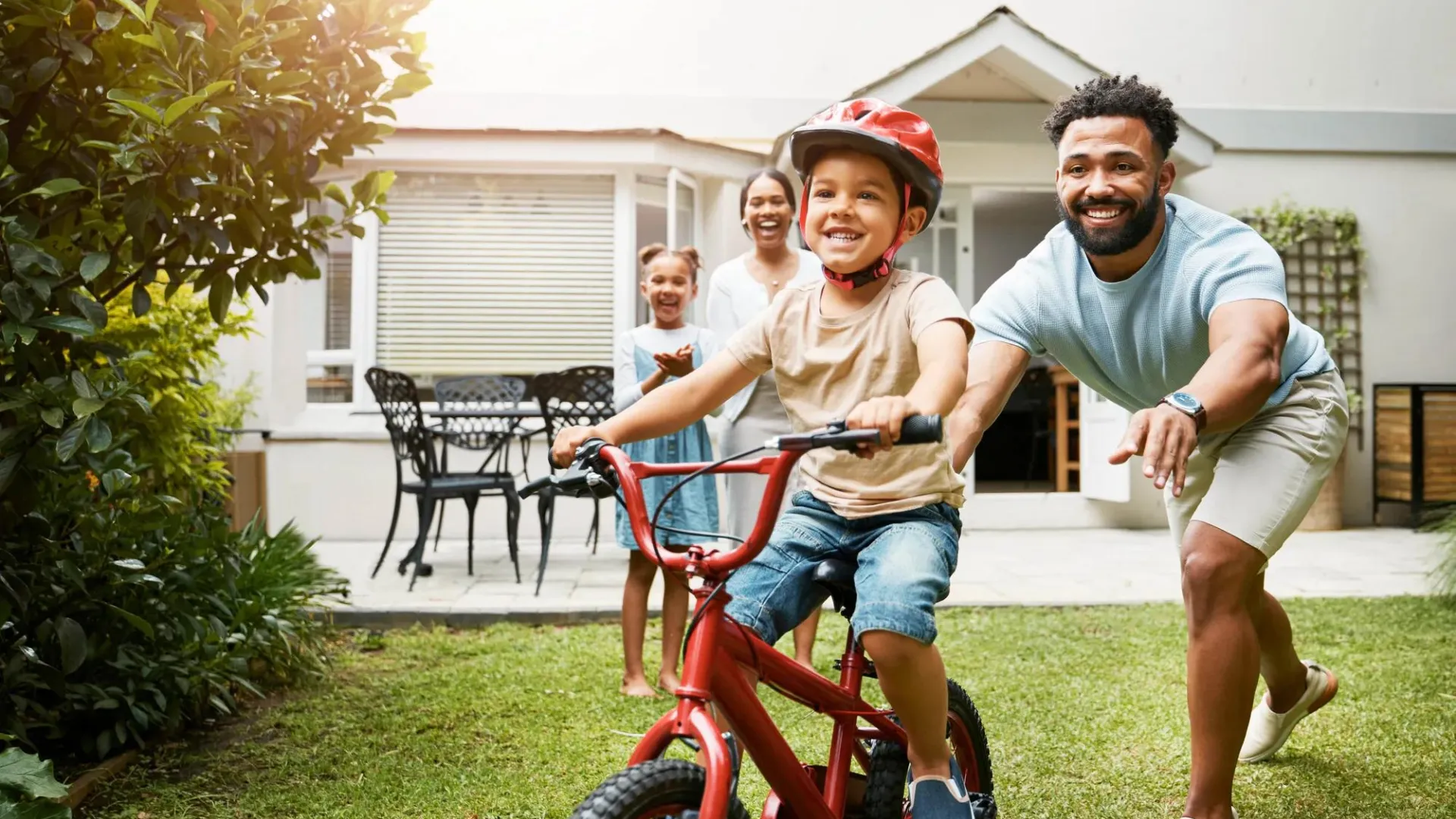 Two parents and two children playing in backyard with one child learning to ride a bike.