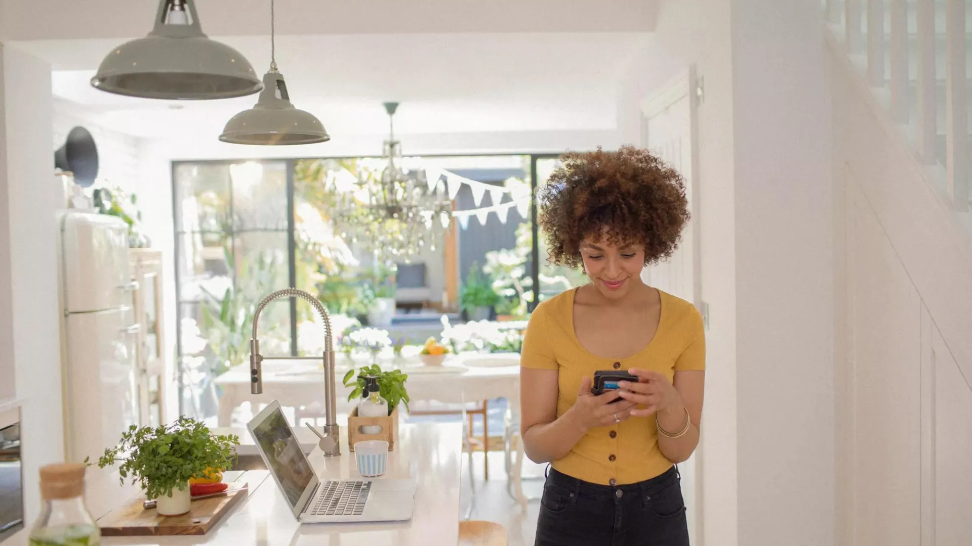 Young woman using smart phone and computer to check health savings account standing in sunny kitchen.