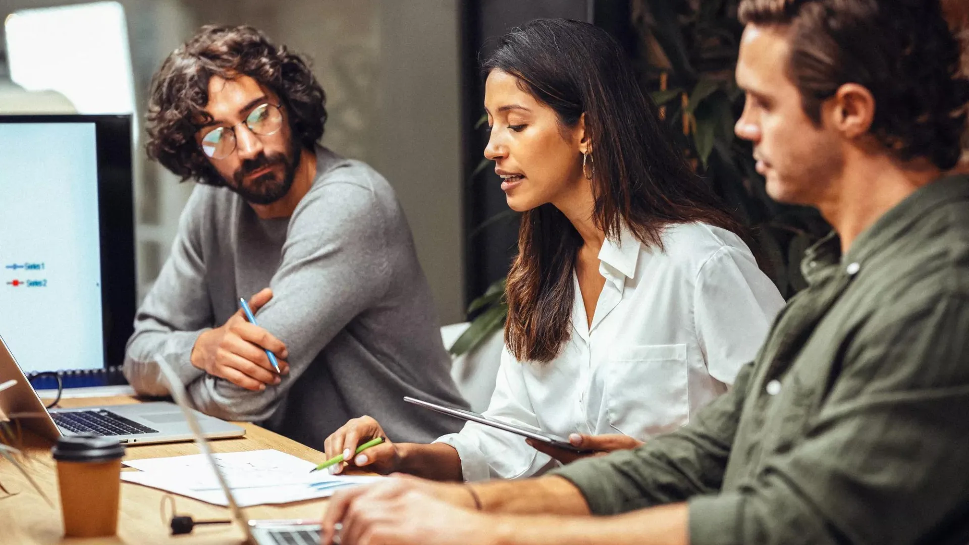 Three employees collaborating at a desk looking at their computers and documents.
