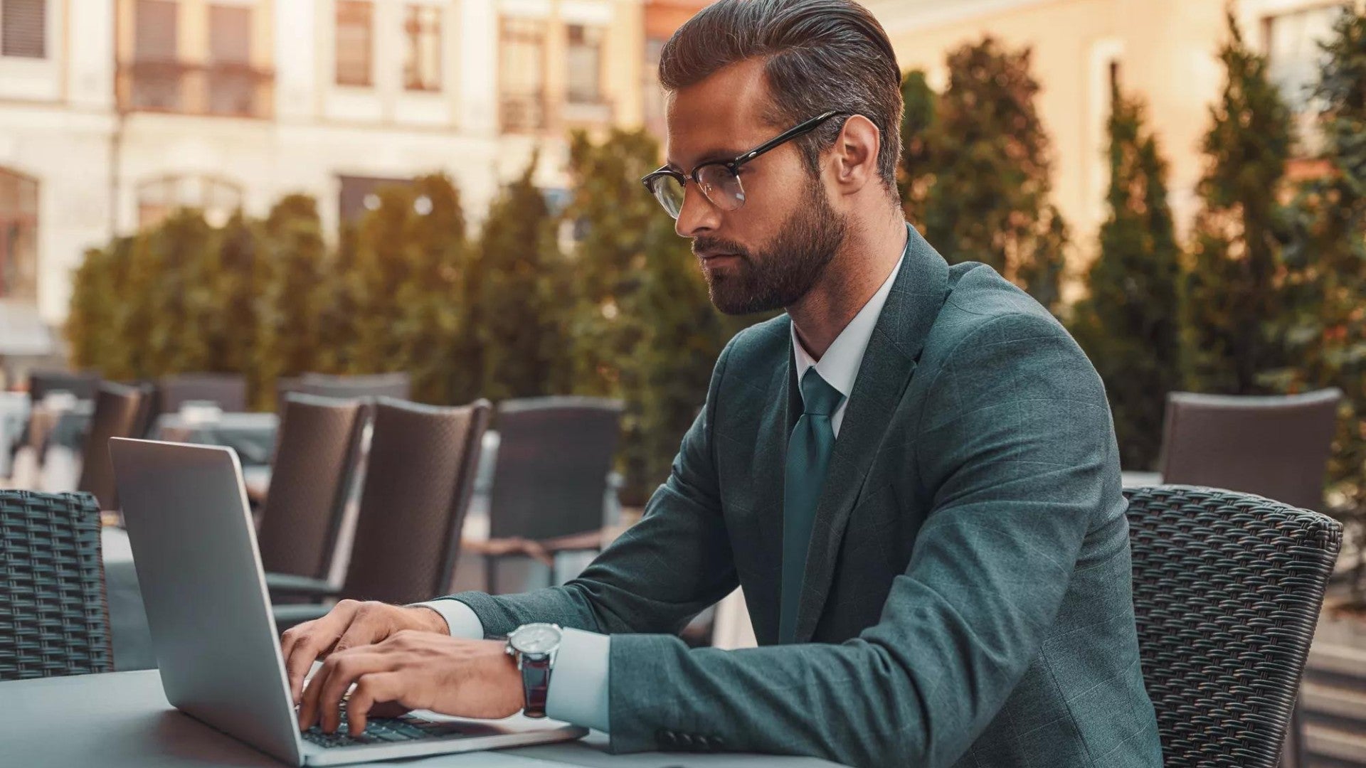 Portrait of bearded businessman in eyeglasses working with laptop while sitting in restaurant outdoors.