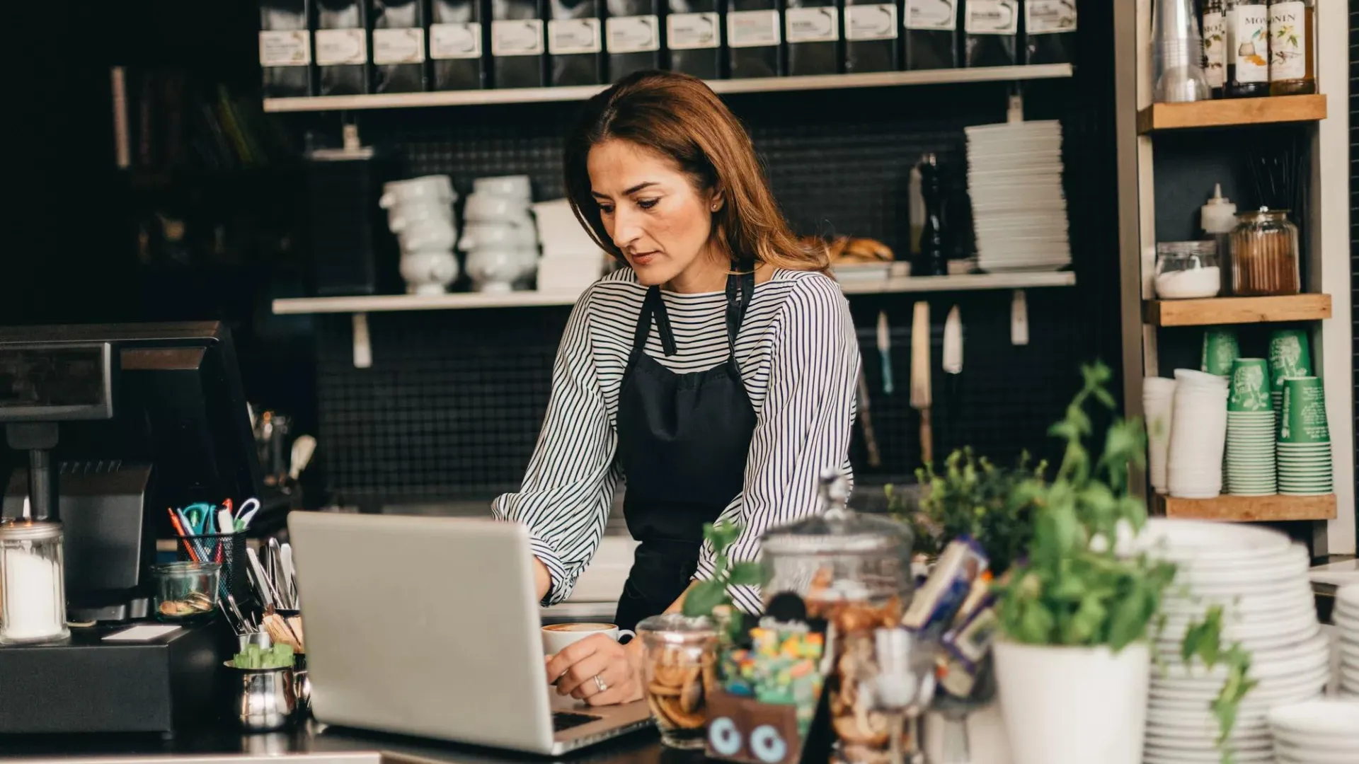 A café owner stands at the counter working on her laptop