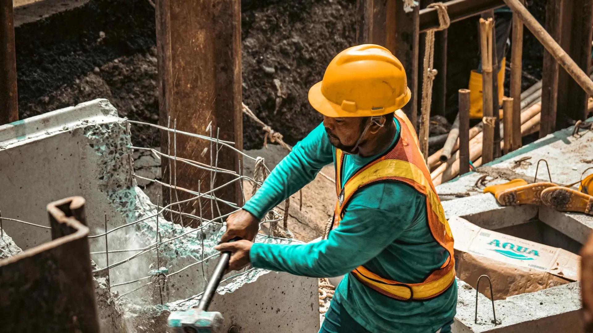 Worker in a hardhat and orange safety vest using a sledgehammer to break up a piece of concrete.