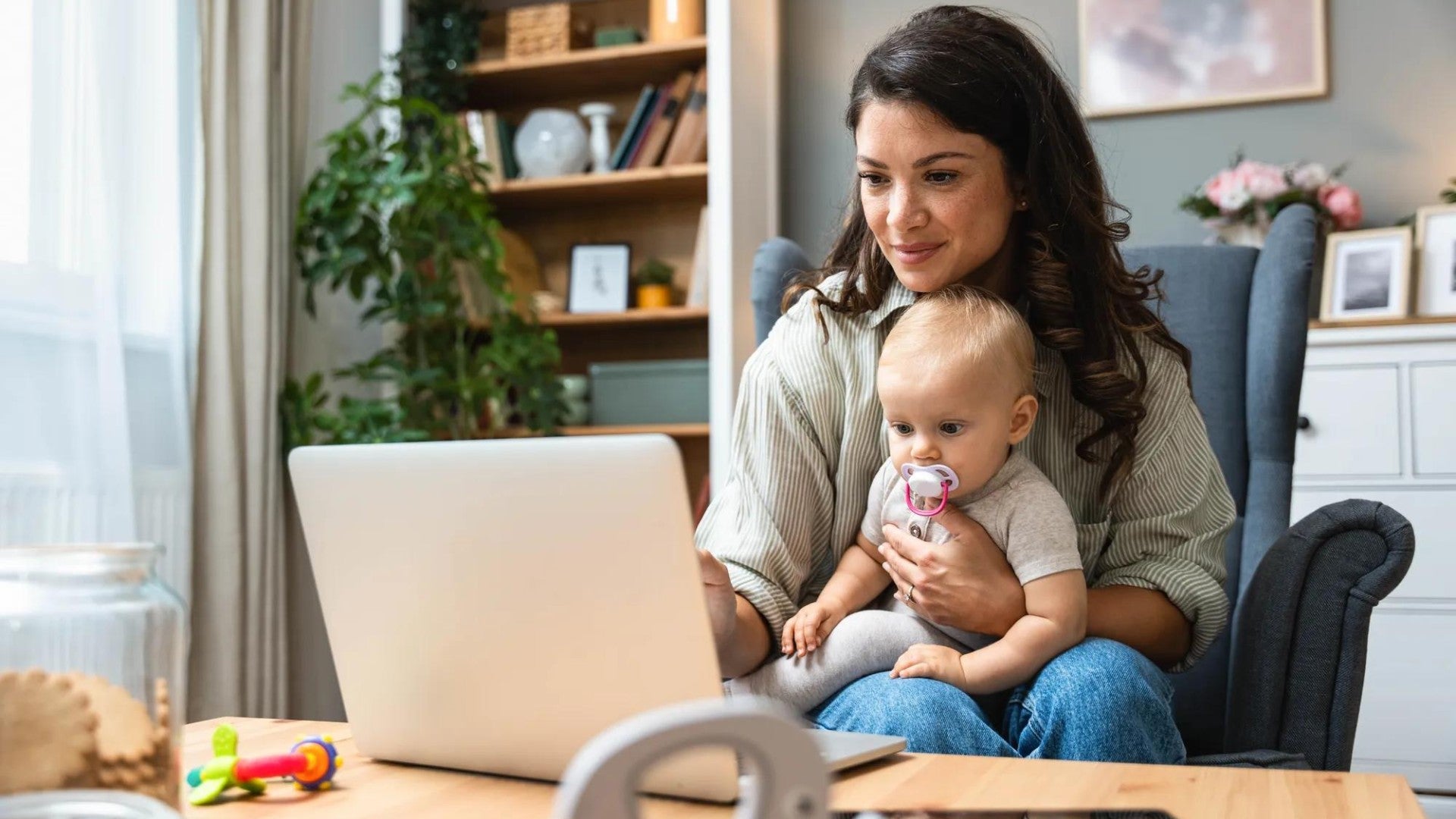 Woman sitting at her dining room table holding her infant while working on her laptop. Woman sitting at her dining room table holding her infant while working on her laptop.