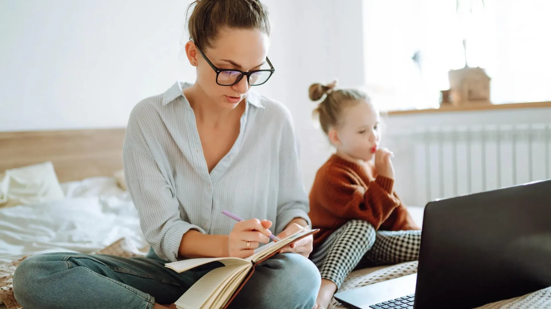 Mother and her young daughter sitting on a bed with a notebook in hand and laptop open.
