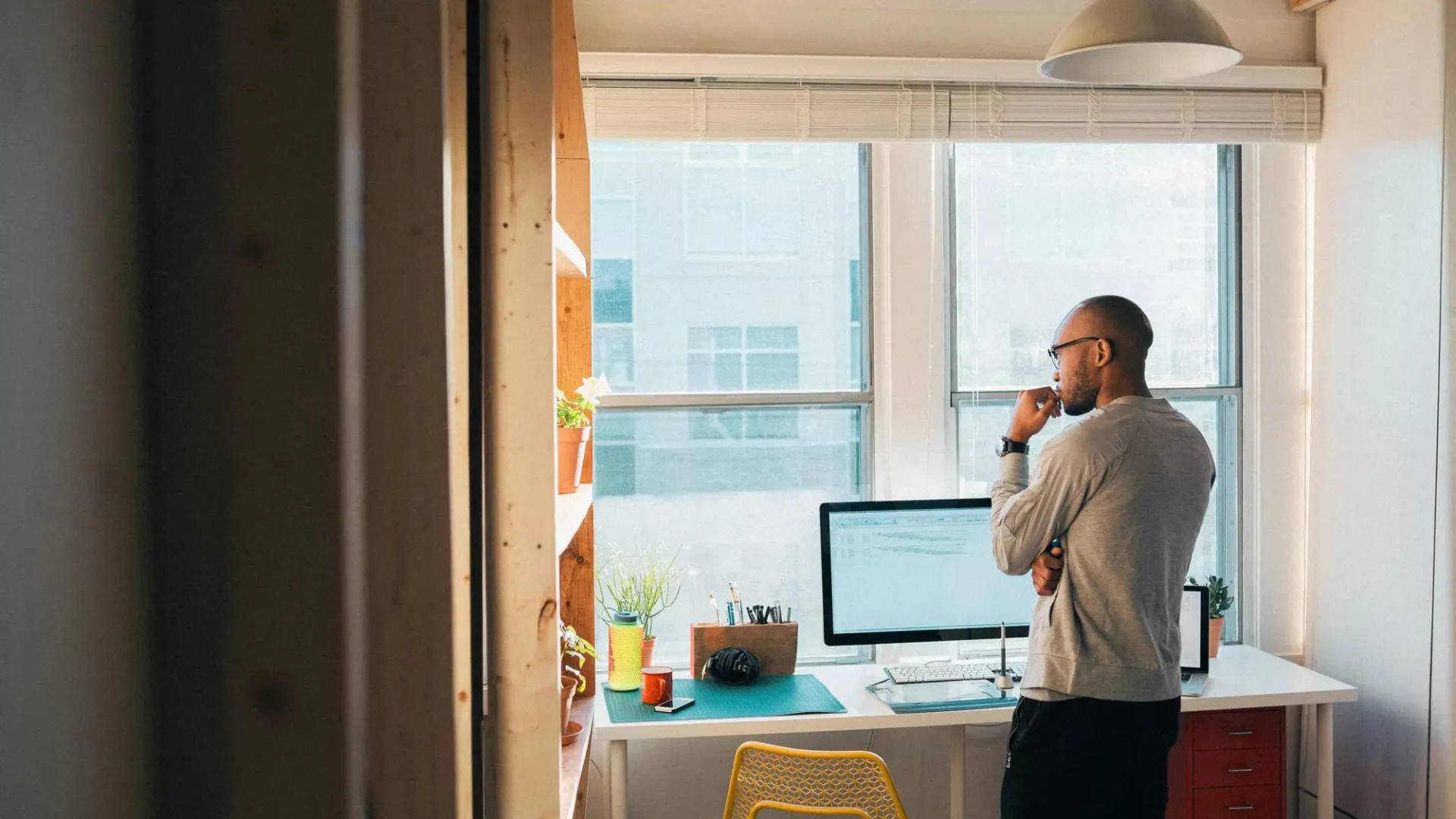 A man standing in a modern, sunlit home office, looking thoughtfully at a computer screen.