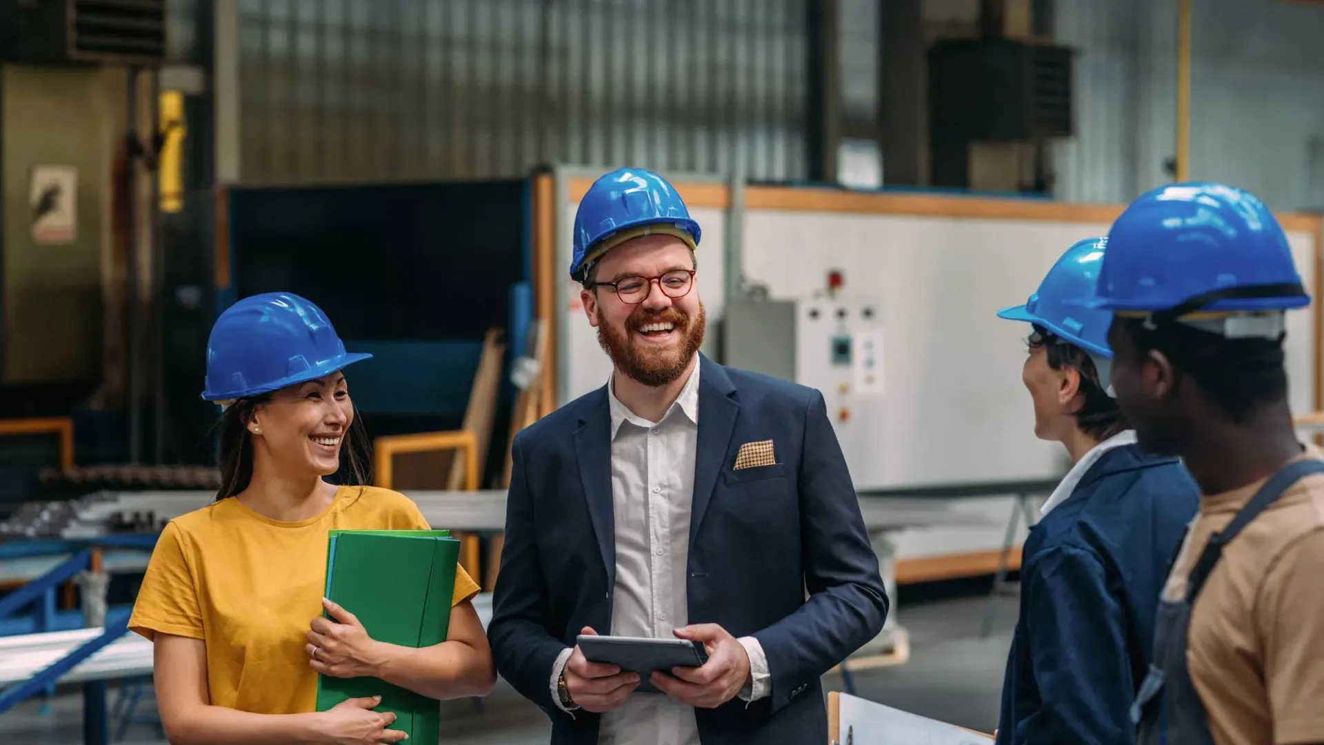 A day in the factory, workers and managers with tablets and clipboards in hard hats smiling in conversation.