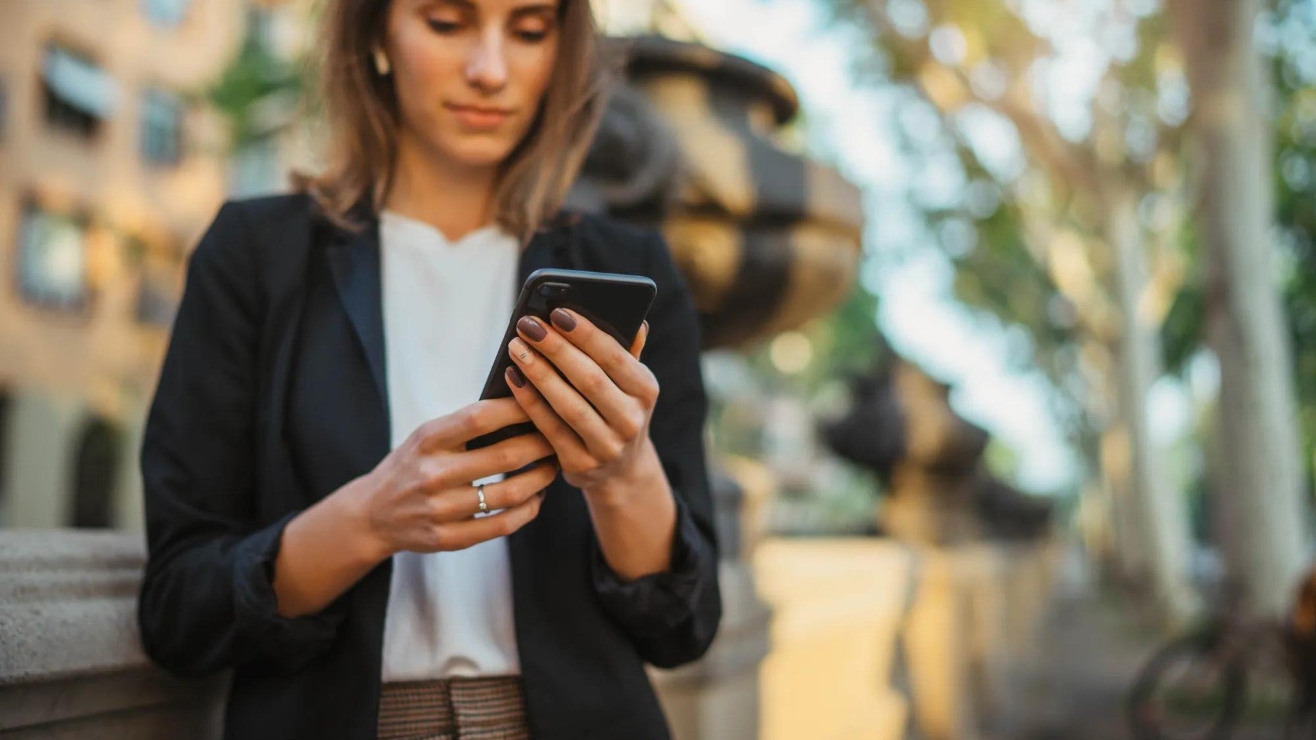 A person in professional attire wearing a black blazer and white top checks their mobile phone while standing outdoors