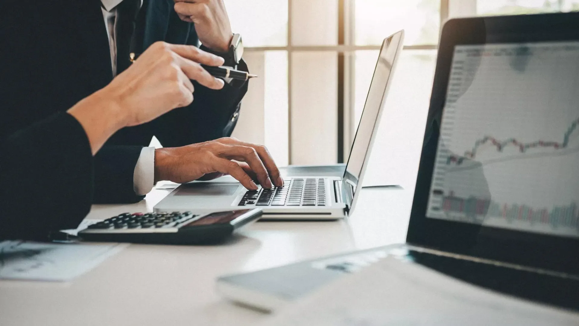 Business professional typing on laptop at desk with financial charts visible, photographed from side angle in natural light.