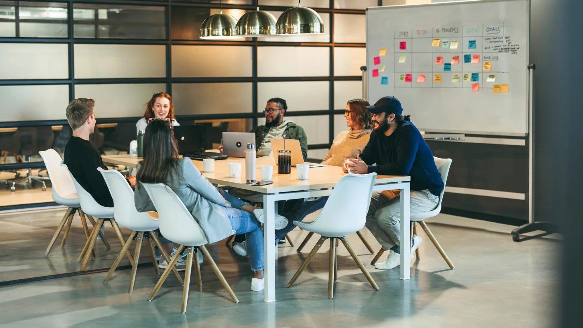 Six employees around a desk having a brainstorming session in front of a whiteboard with sticky notes.