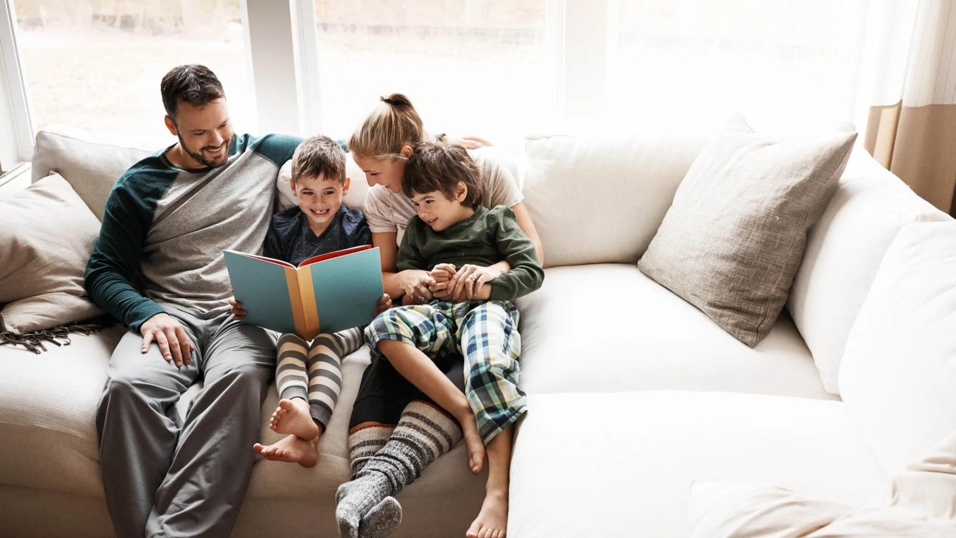 Family snuggled together on white couch reading a book, wearing casual clothes and cozy socks in natural lighting.