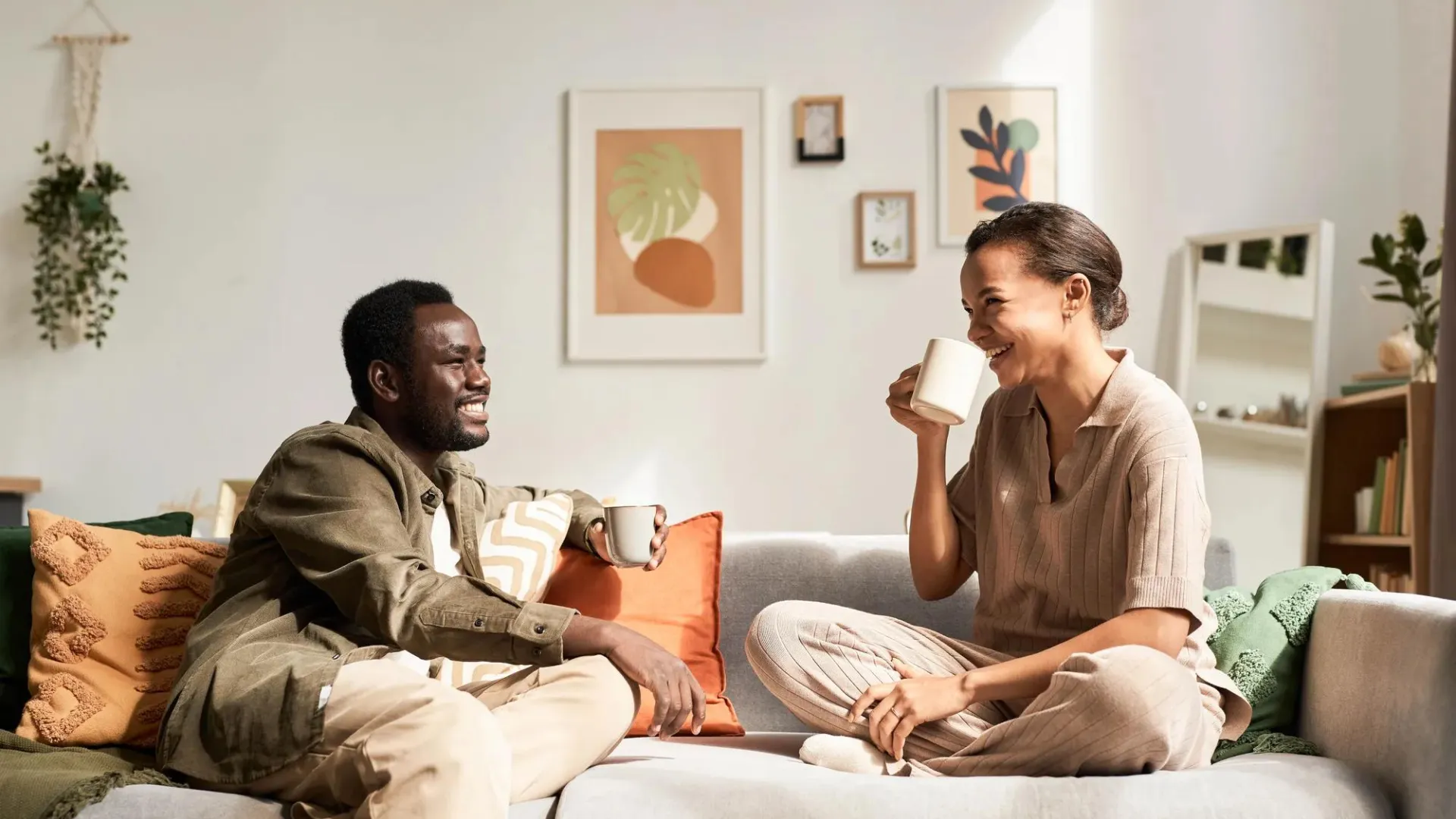 Two people relaxing on a couch, sharing coffee and laughing in a bright living room with modern wall art and plants