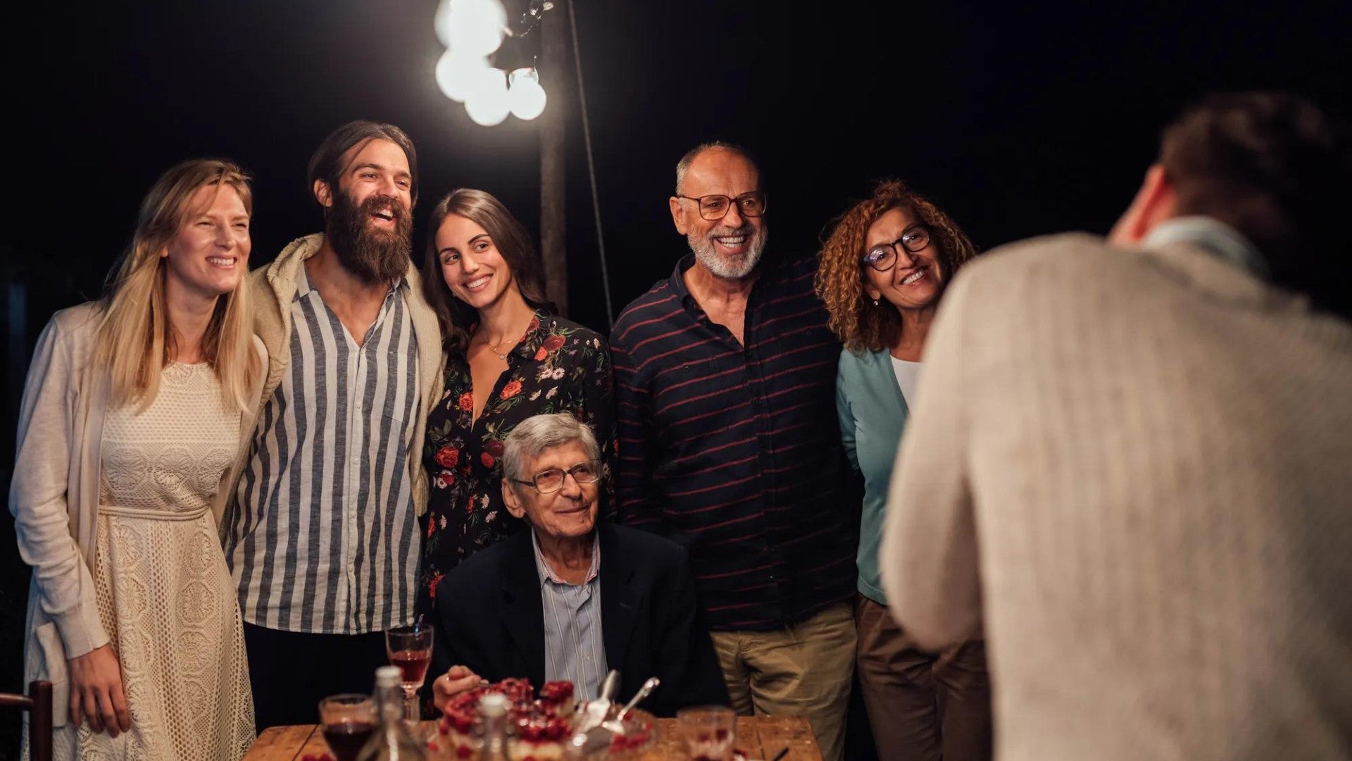 A group of people smiling at an evening gathering around a table with desserts, under string lights. Casual, warm atmosphere.