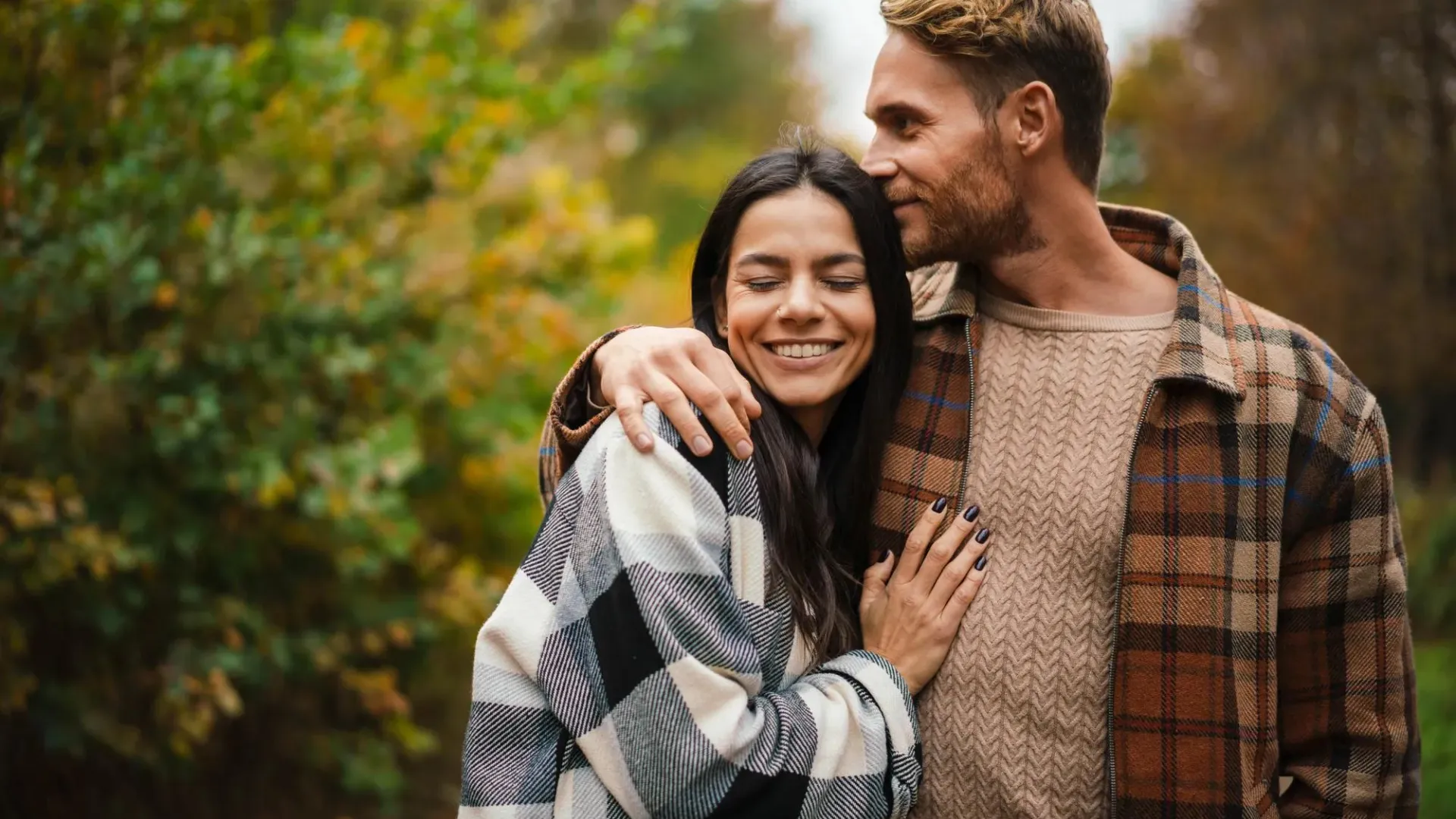 A couple embracing outdoors in fall, wearing cozy plaid and knit sweaters. Autumn foliage in background.