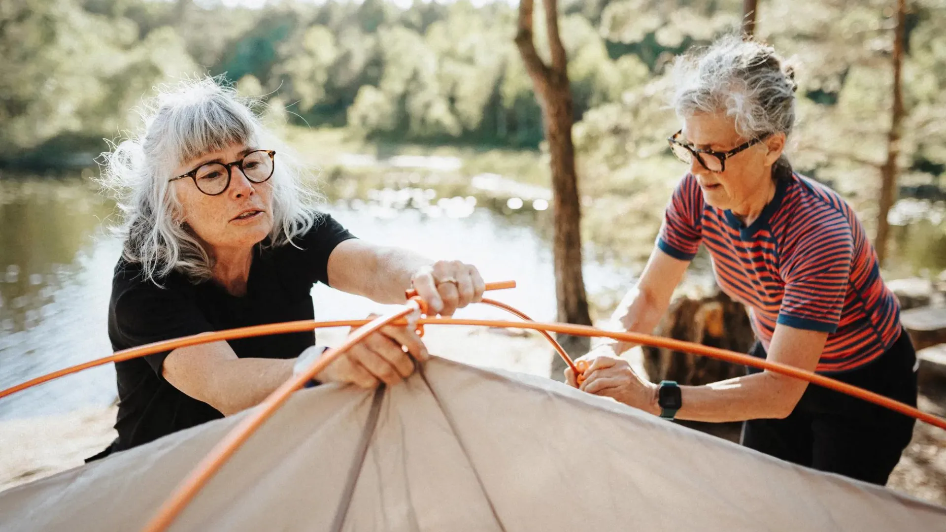 Two retirement age women setting up a tent together beside a lake in a sunny forested camping area.
