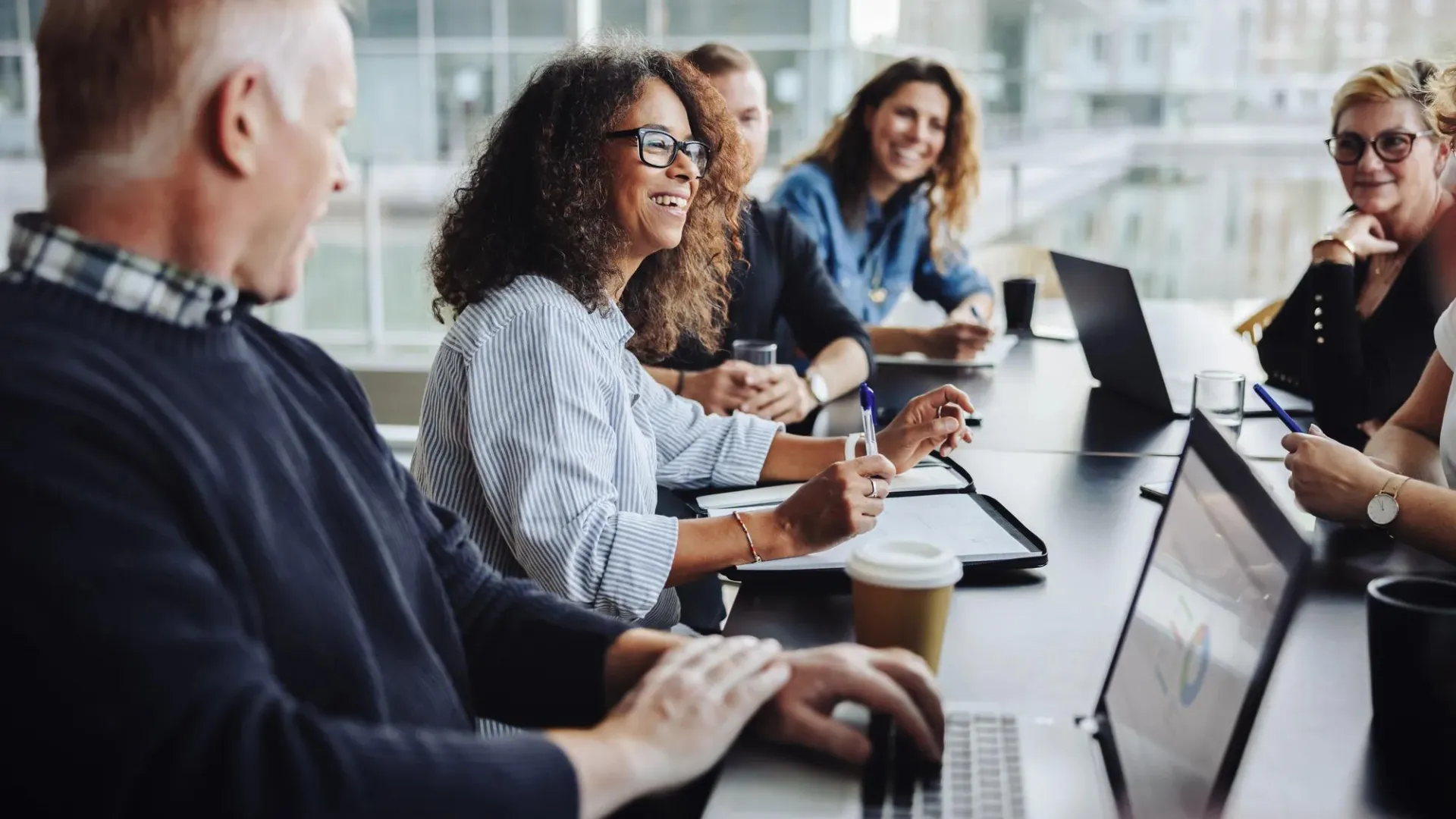Diverse group of professionals collaborate in bright office setting, sharing ideas over laptops with coffee in a casual meeting.