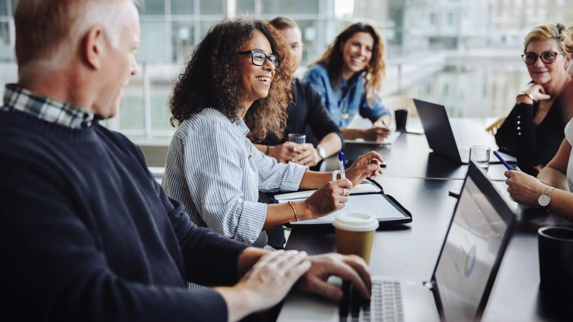 Diverse group of professionals collaborate in bright office setting, sharing ideas over laptops with coffee in a casual meeting.