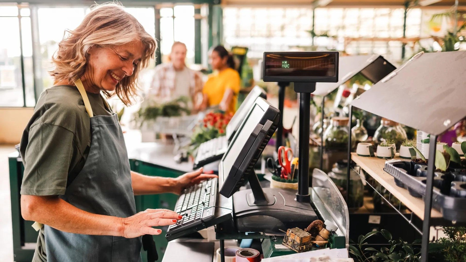 A florist smiles as she operates the cash register in her greenhouse, with customers in the background.