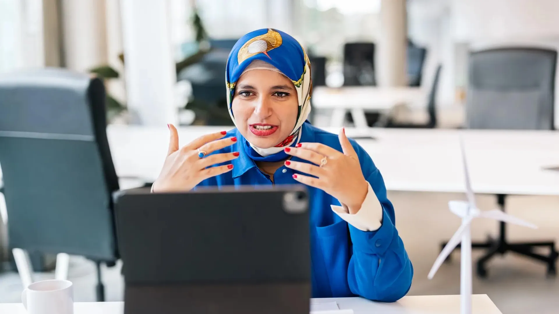 A businesswoman wearing a hijab conducts a videoconference on her laptop computer while sitting in her office. 