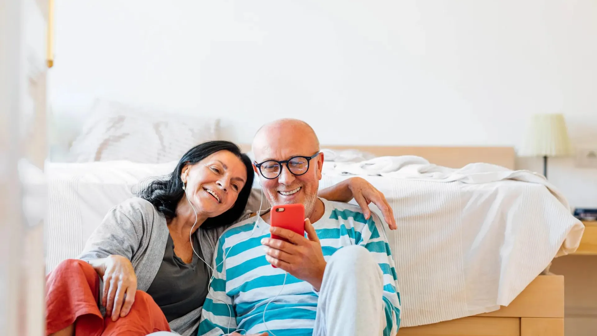 Man and woman sitting in front of a bed listening to music on earbuds 
