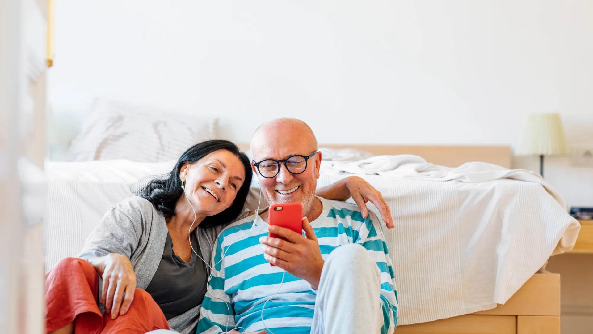 Man and woman sitting in front of a bed listening to music on earbuds 