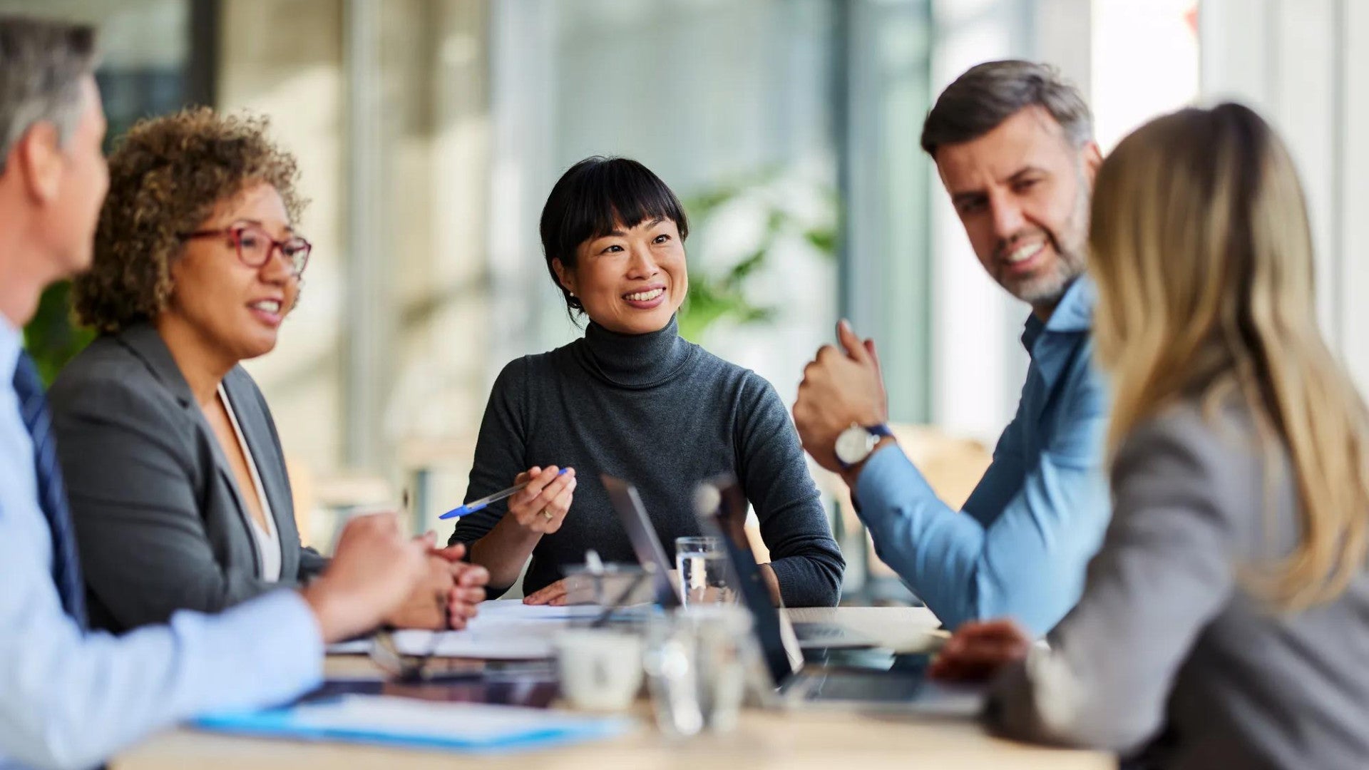 Group of professionals seated around a table in an office, engaged in discussion with documents, pens, and laptops visible.