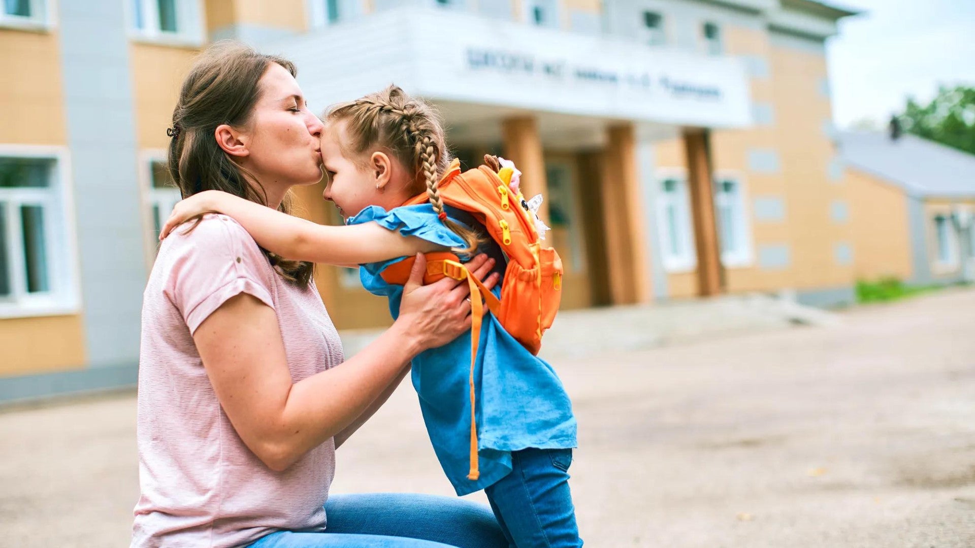 Mom sitting on a curb giving a kiss to daughter wearing a backpack