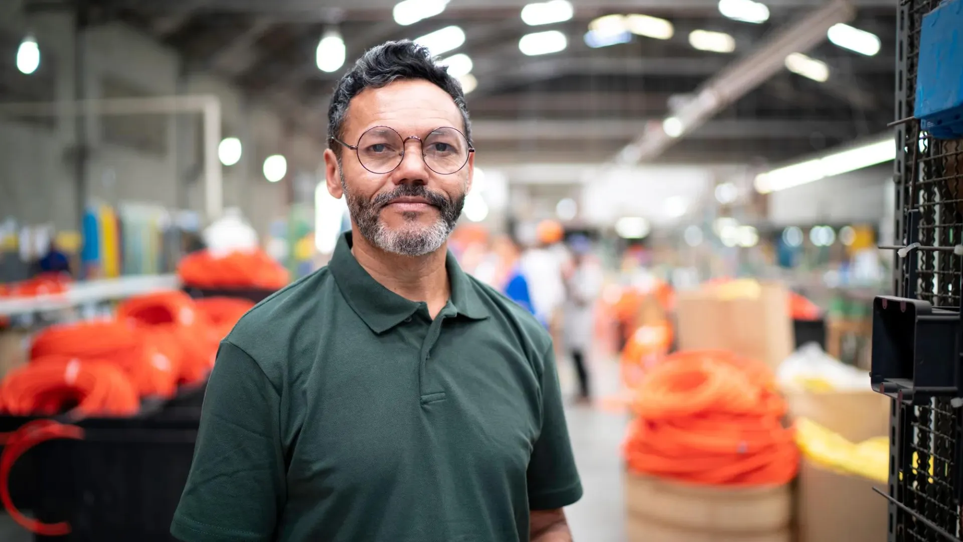 Person wearing a green shirt stands in a warehouse with stacked boxes and coiled orange hoses in the background
