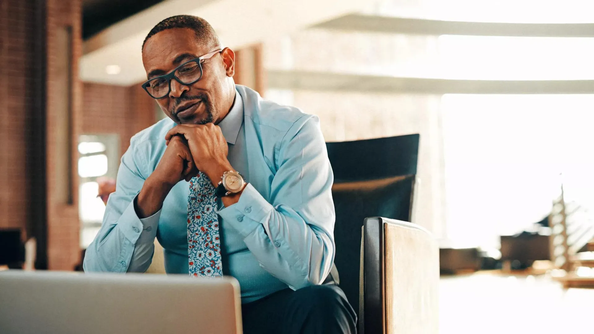Person sitting in office chair, hands clasped, looking at laptop, wearing glasses, dress shirt, and sassy tie.