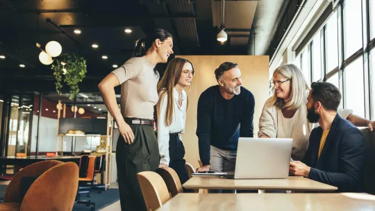 Five colleagues of various ages working together around a laptop in a modern office environment.