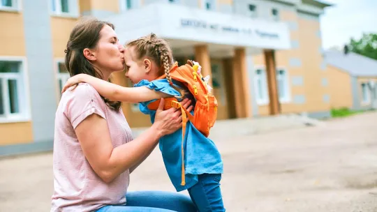 Mom sitting on a curb giving a kiss to daughter wearing a backpack