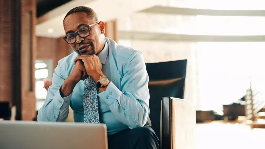 Person sitting in office chair, hands clasped, looking at laptop, wearing glasses, dress shirt, and sassy tie.