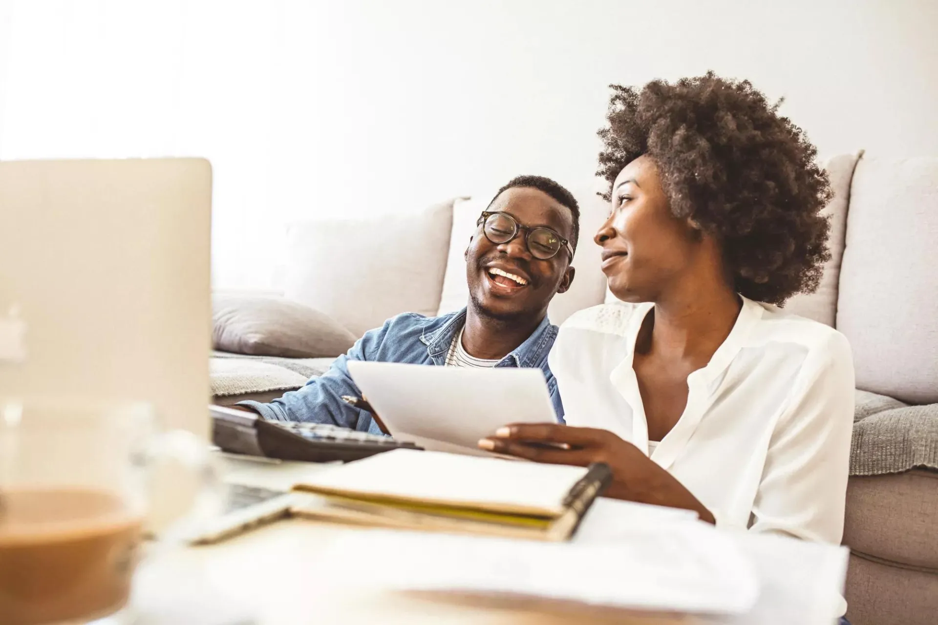 A young couple smiles while reviewing financial documents together on their couch at home.