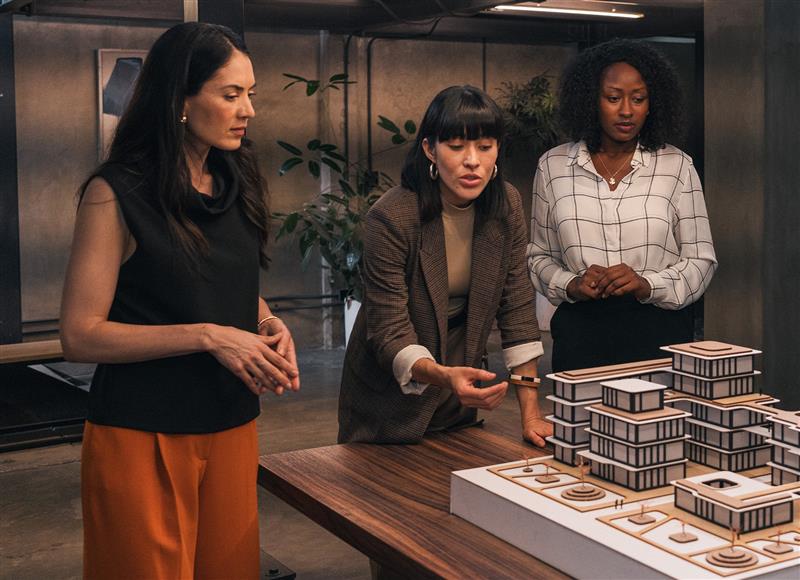 Three businesswomen professionals discuss how to solve a problem while gesturing toward a building diorama