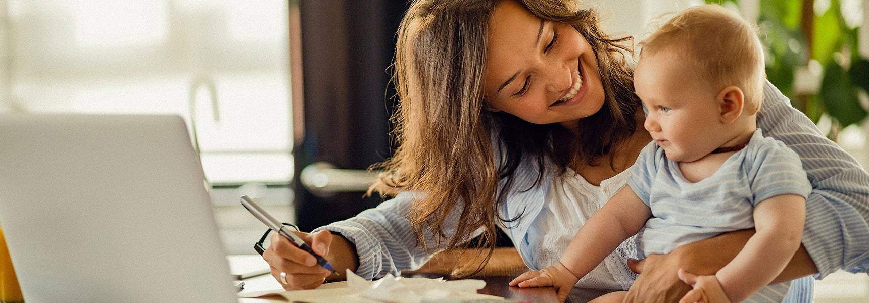 Photo of a woman smiling at baby while she works on a laptop.