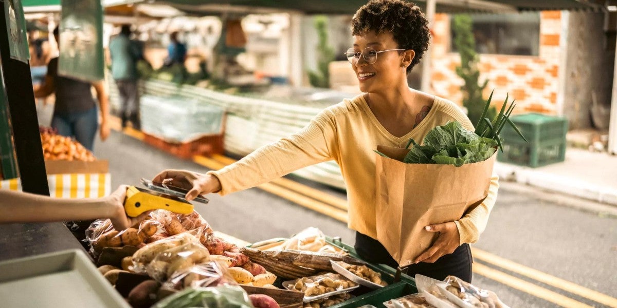 Woman at an outdoor farmer’s market uses her phone to tap to pay the vendor for her bag of produce