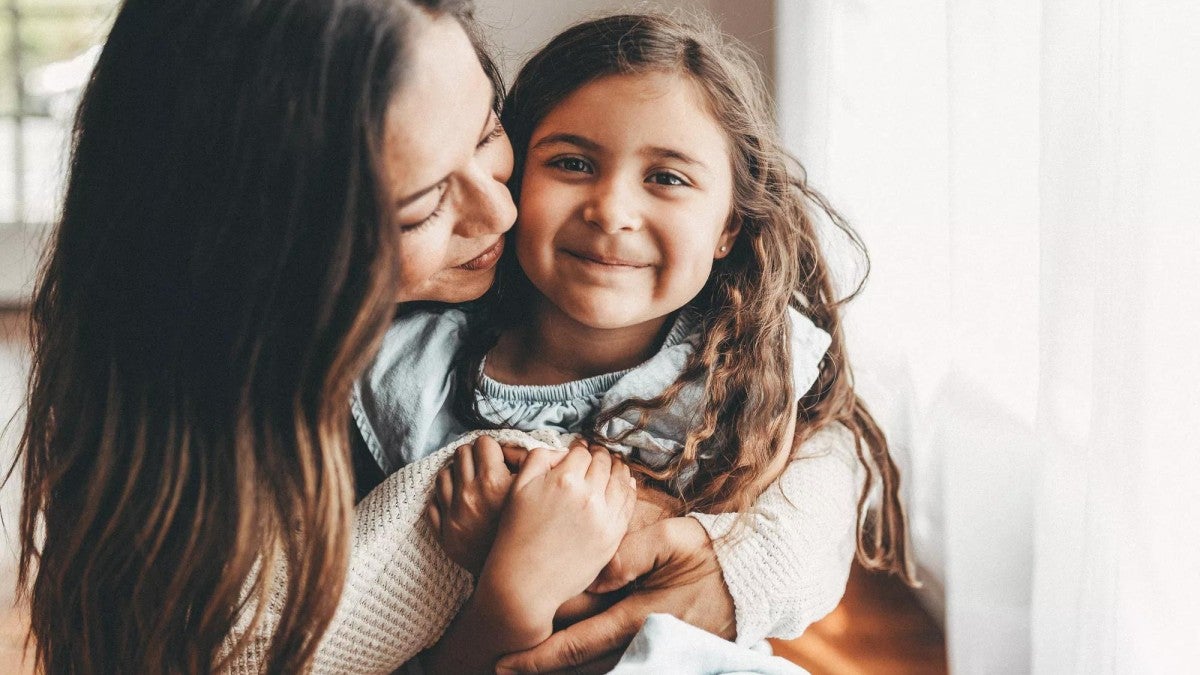 Woman wraps her arms around her young, smiling daughter.