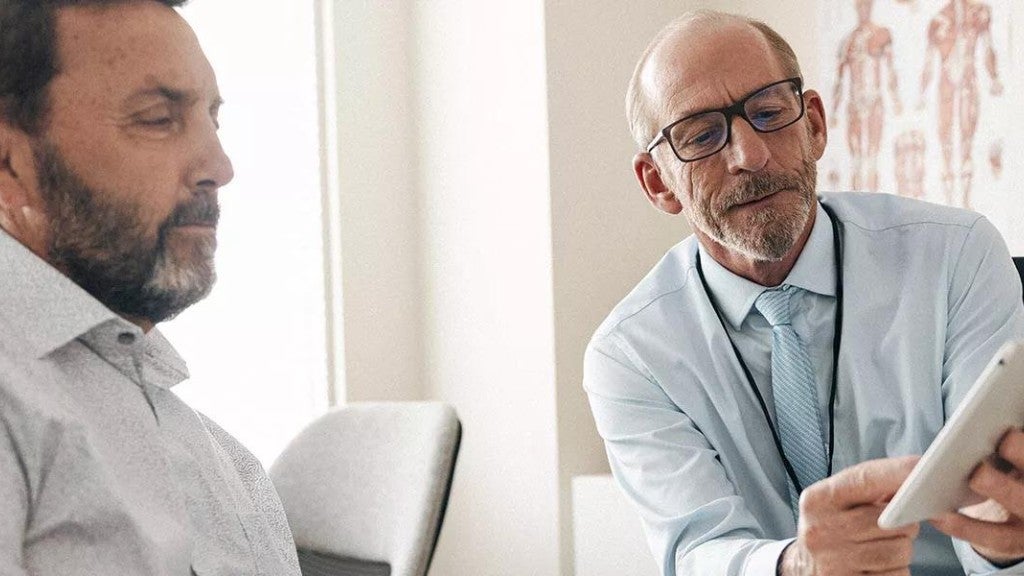 A patient in a doctor's office looks at a tablet a doctor is holding while the doctor explains what he is seeing.