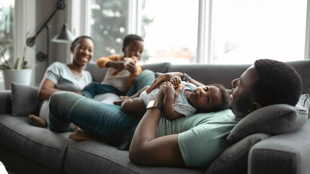 Happy Black family relaxing on a  gray couch with man holding a baby and woman next to a toddler.
