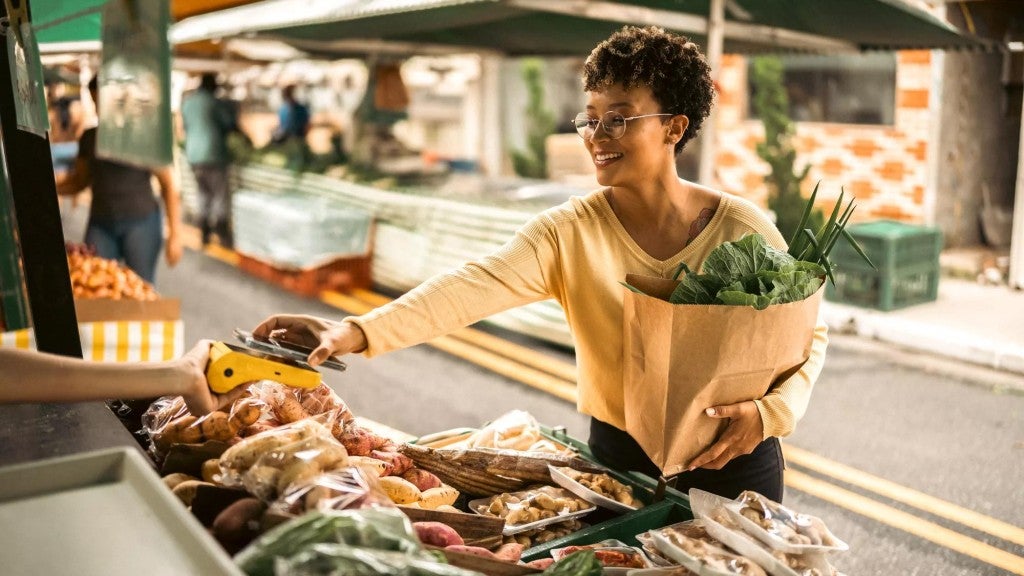 Woman at an outdoor farmer’s market uses her phone to tap to pay the vendor for her bag of produce