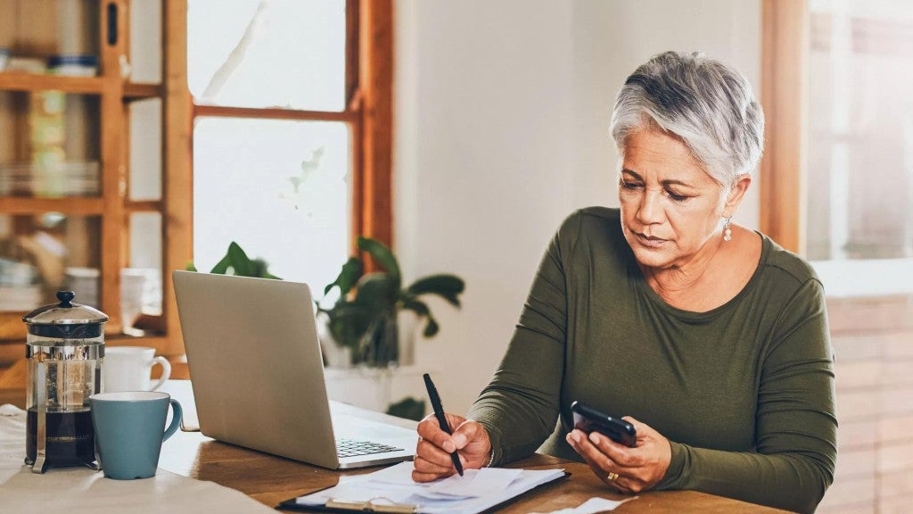 Women working at a table with her laptop and cell phone, writing on paper. 