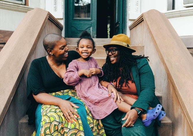 Two women smiling on a porch stoop while looking at a young daughter