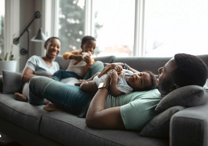 Happy Black family relaxing on a  gray couch with man holding a baby and woman next to a toddler.