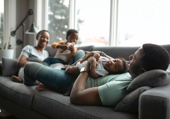 Happy Black family relaxing on a gray couch with man holding a baby and woman next to a toddler.