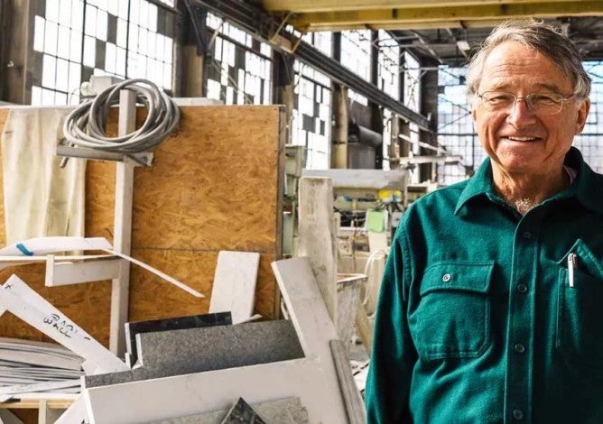 An older entrepreneur stands smiling in a brightly light production facility 