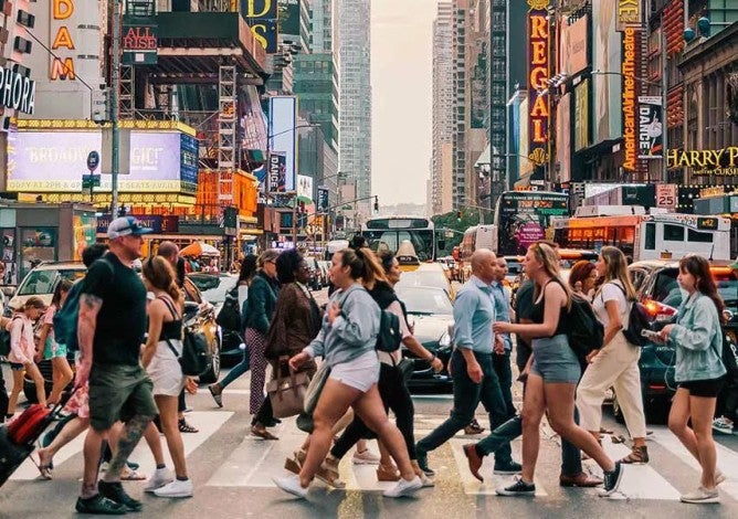 Photo of dozens of people walking across a crowded crosswalk at a busy city intersection.