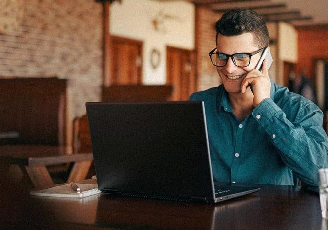 Photo of a man on a computer talking on his cell phone.